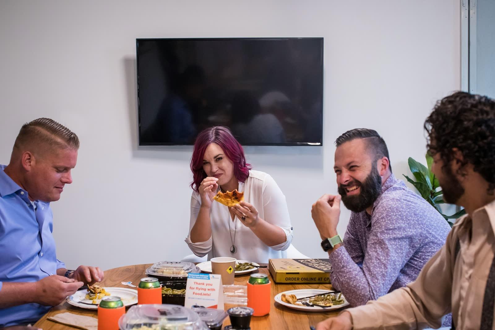 Colleagues laughing around a table in an office while sharing food together