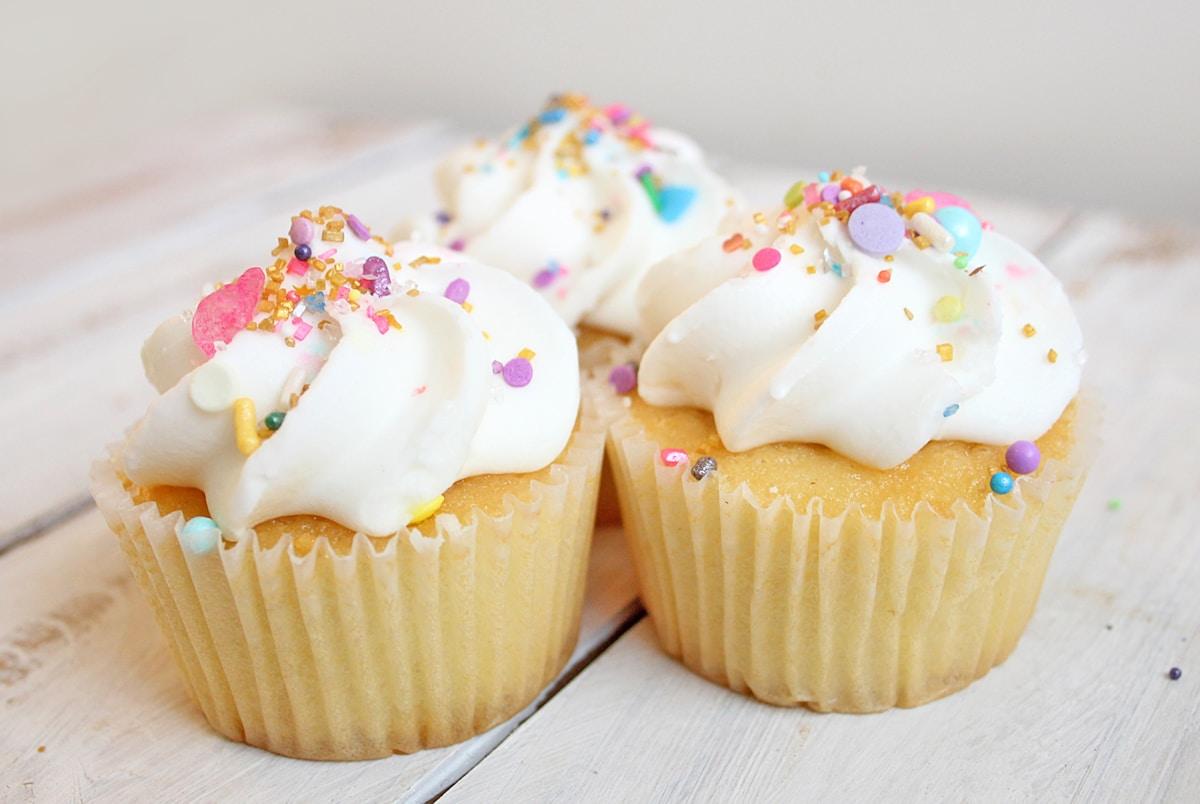 Cupcake alla vaniglia con frosting bianco e codette colorate pastello su una superficie di legno bianco